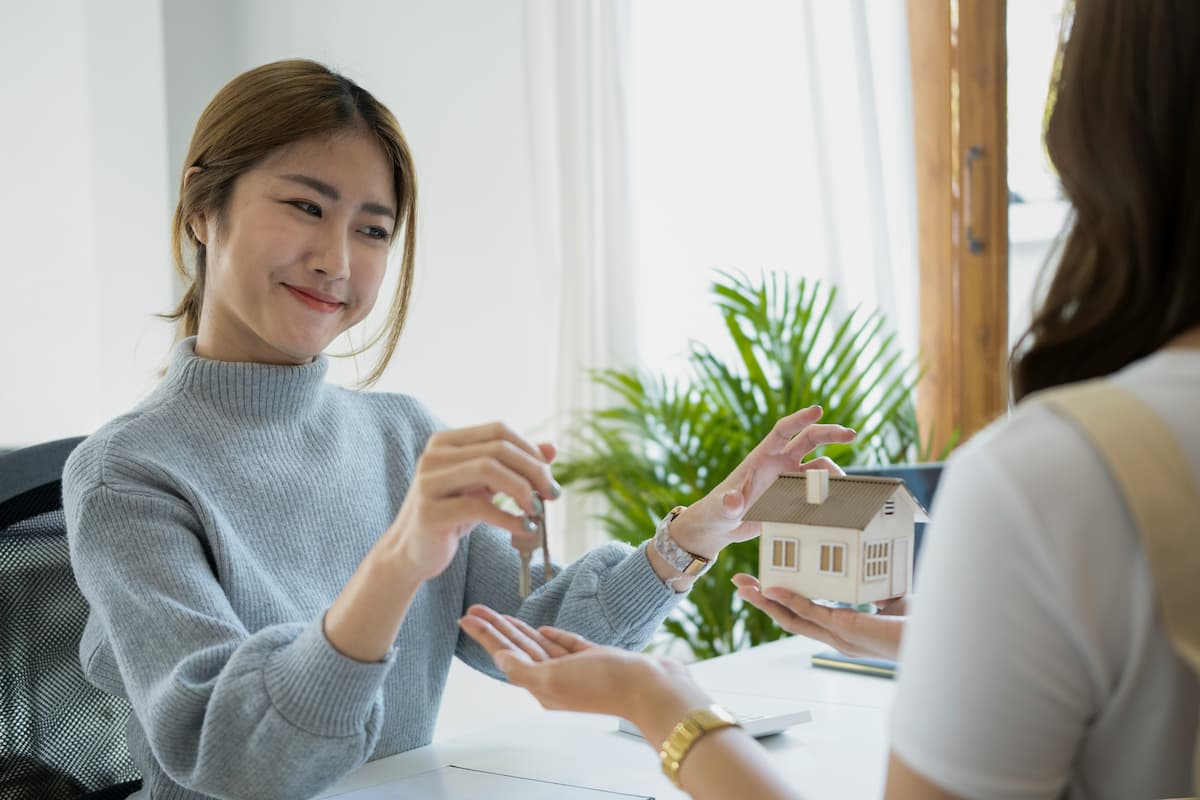 A smiling real estate agent hands a set of keys to a client who is holding a small model house.